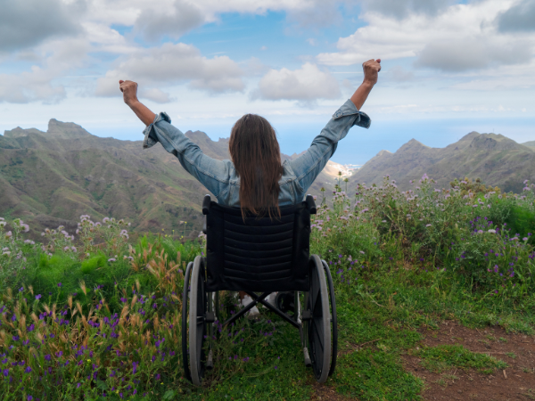 A woman in a wheelchair facing away from the camera with arms outstretched as she looks at scenery