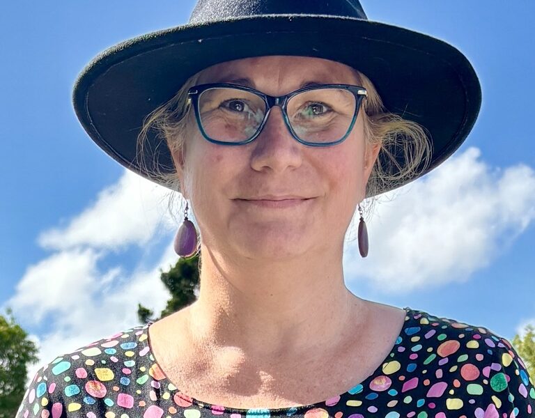 A picture of Sam wearing a black hat, blue glasses and a colourful polkadot dress